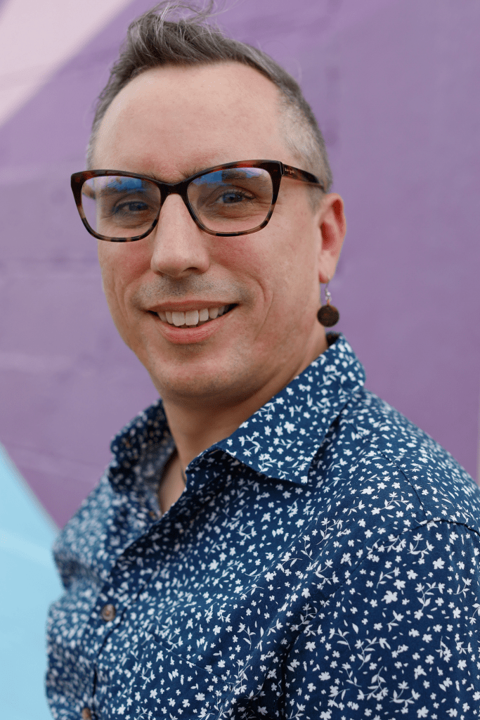 A person wearing a blue collared button-up shirt with small white flowers printed on it while standing in front of a wall with a purple circle and pink and blue backgrounds painted on it.